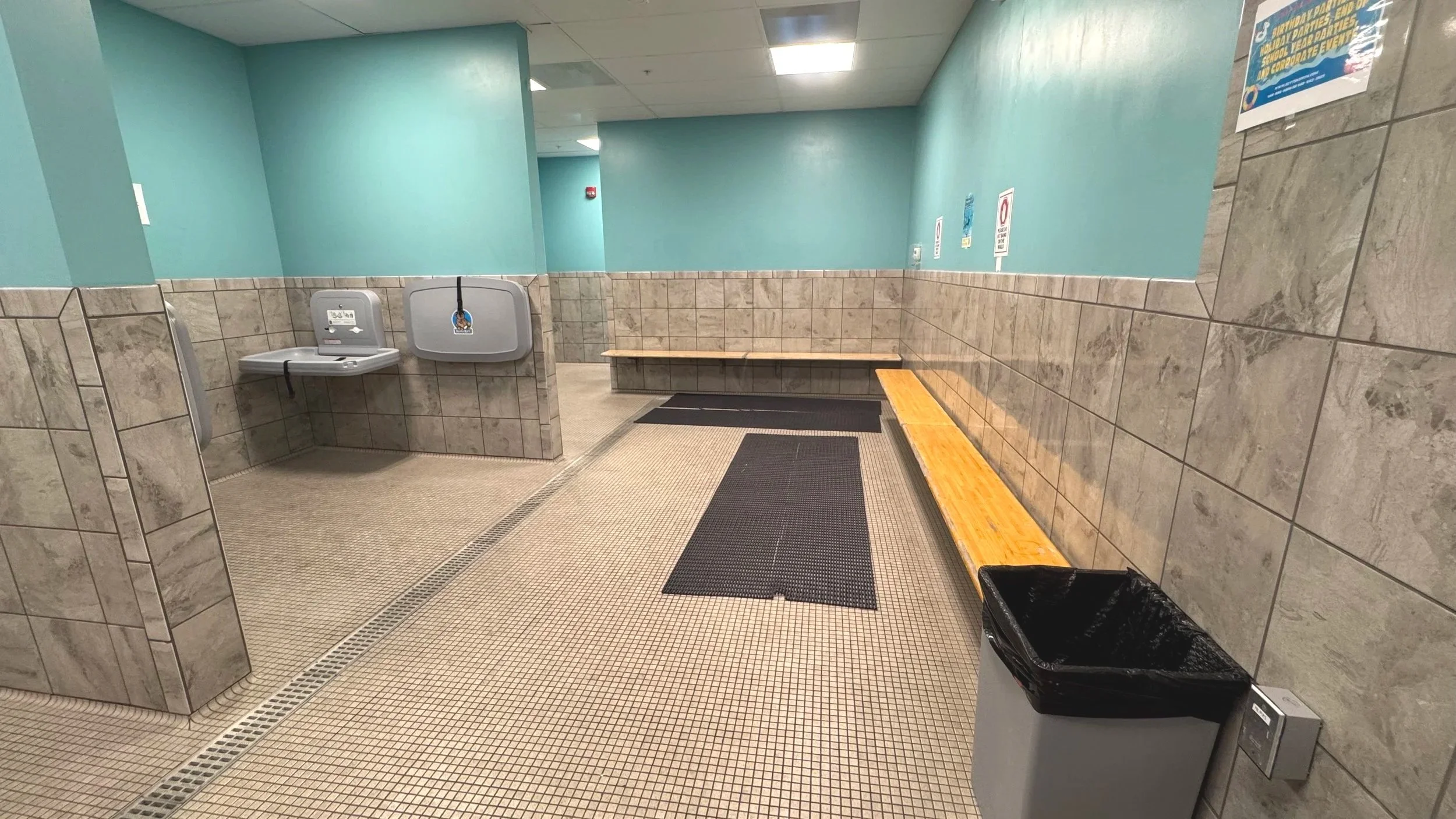 Empty indoor public space with tiled walls and floor, showing water fountains, benches, trash can, and signs, with teal painted walls and ceiling lights.