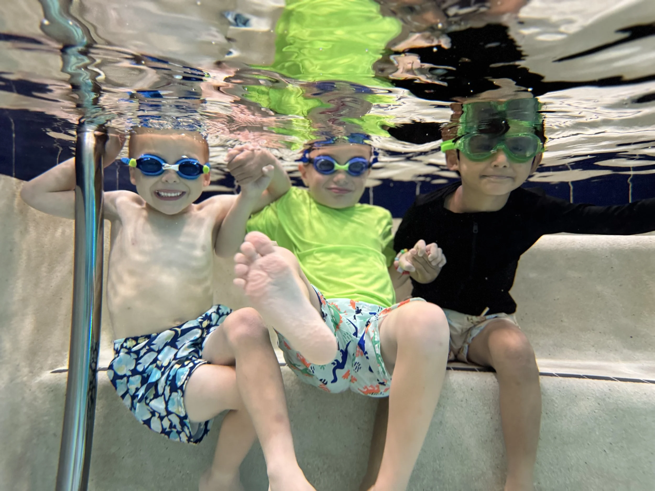 Three children in colorful swim trunks and goggles swim underwater in a pool, smiling and posing for the camera.