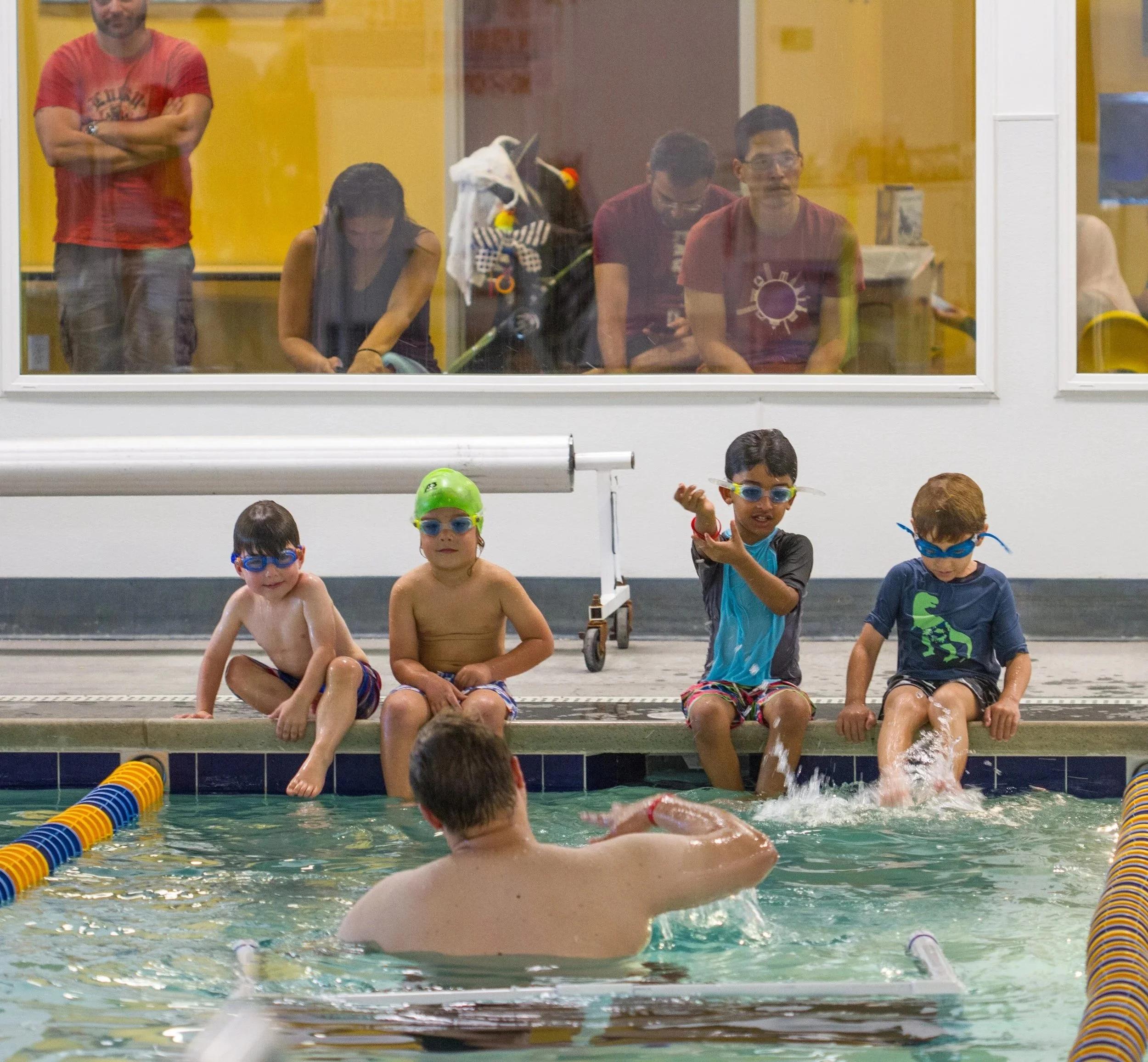 Kids in swimsuits, goggles and swim caps sitting on the edge of a swimming pool, getting ready for their first swim lessons