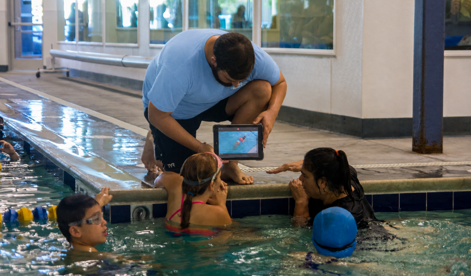 Swim instructor kneeling at the edge of a swimming pool, using video feedback to teach stroke technique to young swimmers