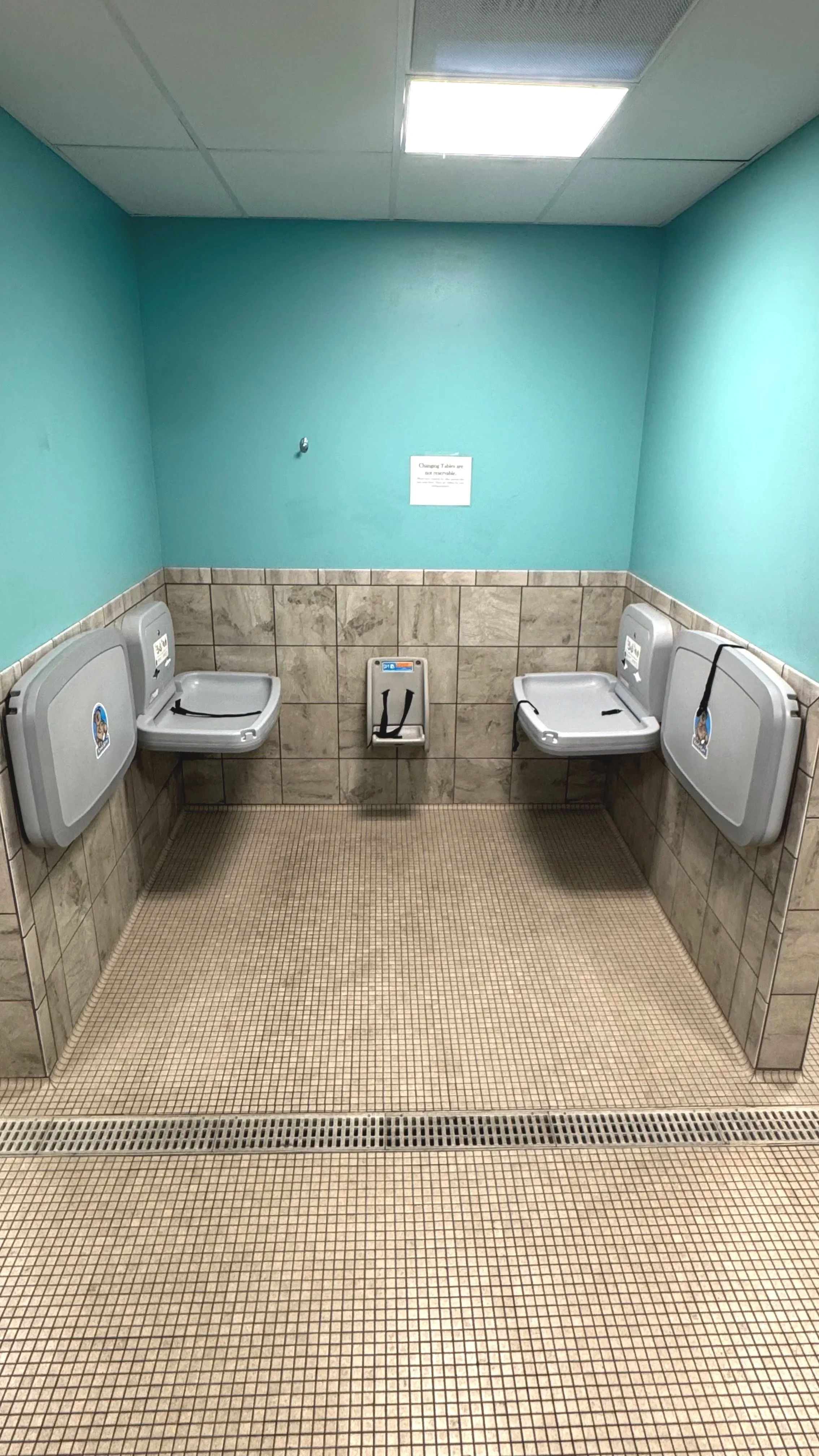 A bathroom with turquoise walls, beige tiled lower wall, beige tiled floor, ceiling with fluorescent lights, and three baby changing stations. Two of them are on the sides and have covers, and one in the center is open with black straps. There is a small sign on the turquoise wall and a small hole possibly for a faucet or flush.