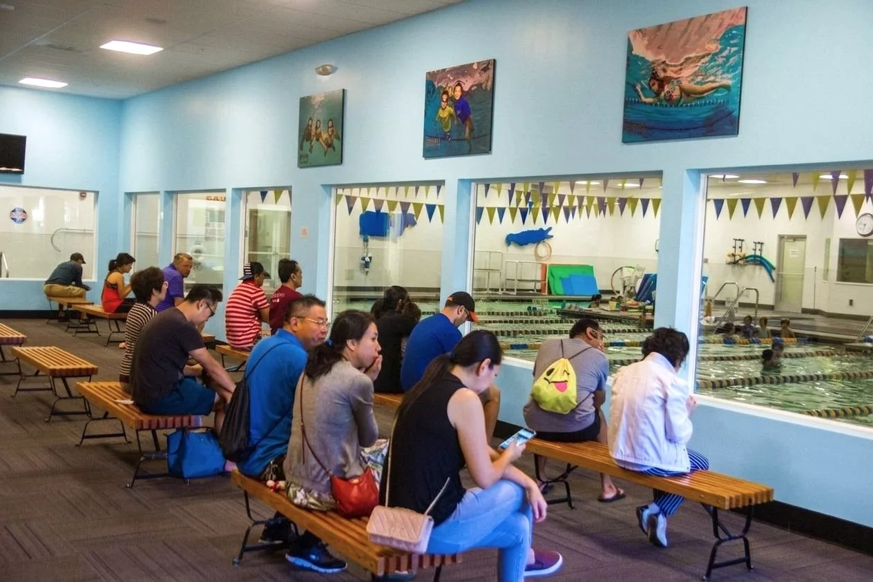 People sitting on wooden benches watching a swimming pool through large windows, with swimmers in the pool and colorful decorations hanging above.