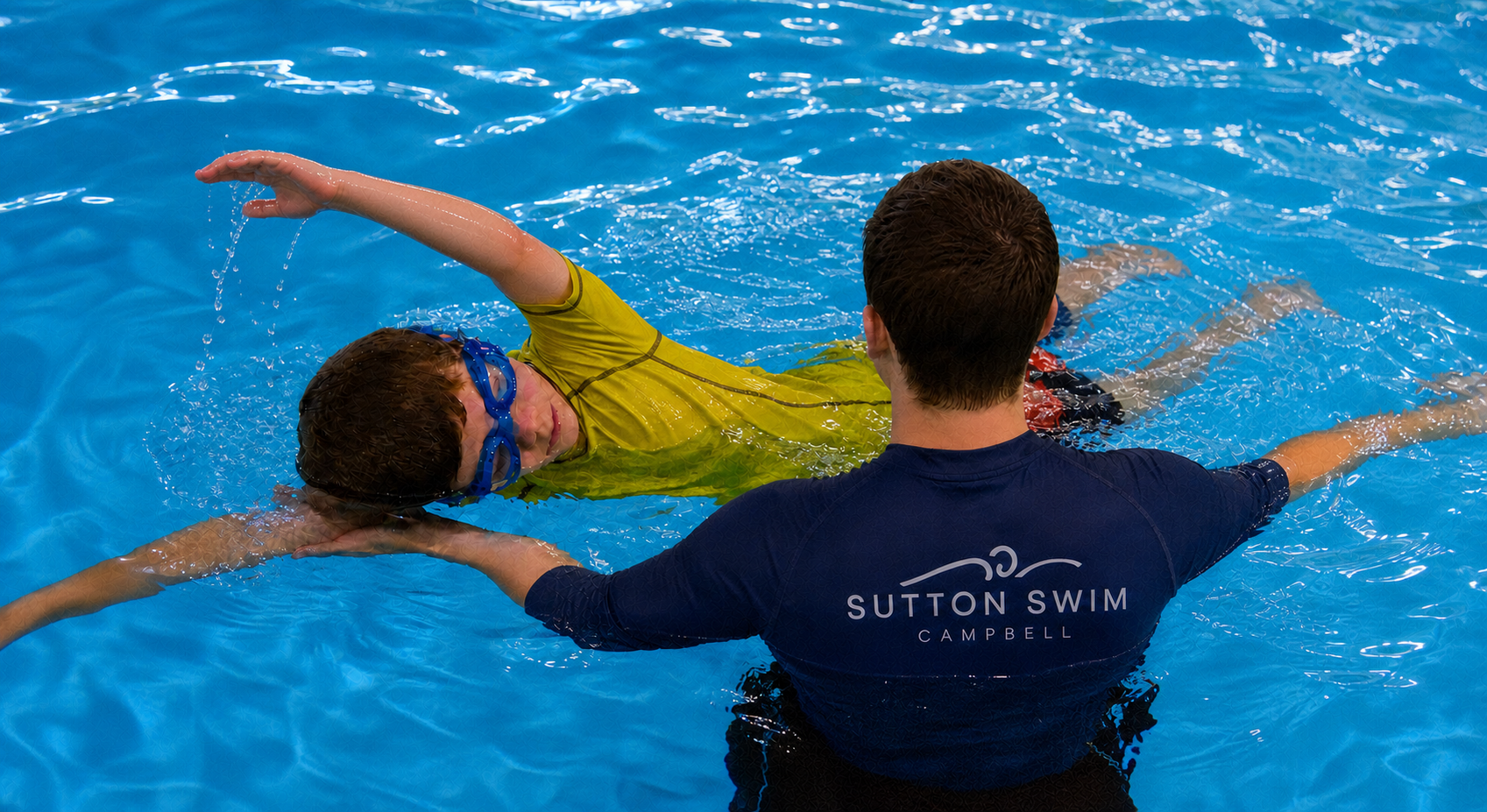 A swim instructor assisting a young boy with swimming in a pool. The boy is wearing yellow swim goggles and a yellow swim shirt, while the instructor is wearing a navy shirt with 'Sutton Swim Campbell' printed on the back. The boy is floating on his 