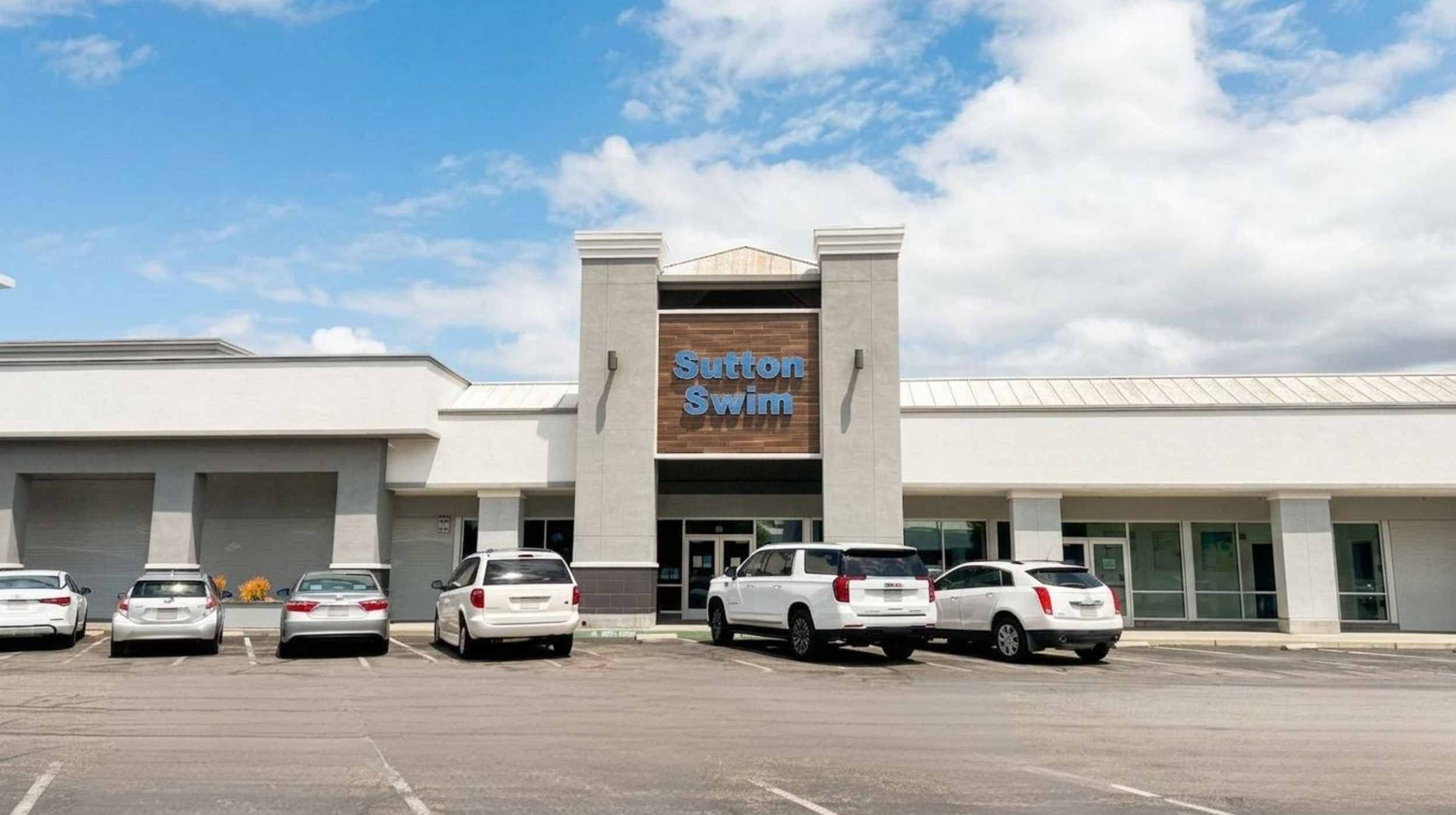 Facade of Sutton Swim with a parking lot in front and several parked cars under a partly cloudy sky.