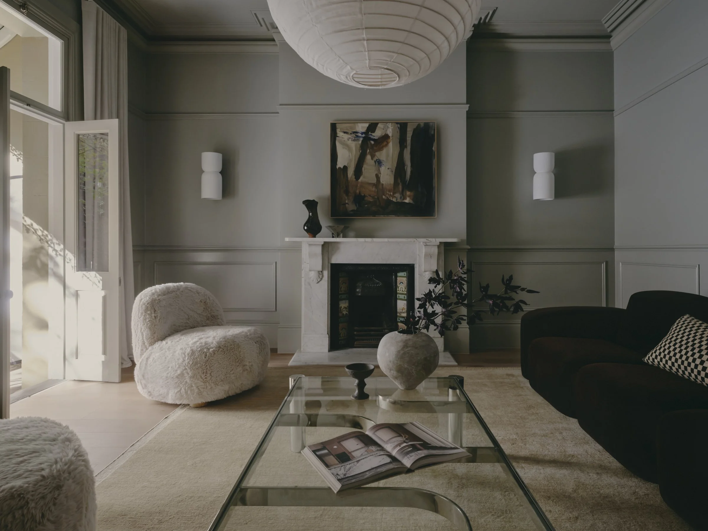 Living room with a marble fireplace, abstract painting above it, white wall sconces, beige rug, cozy beige armchair, black sofa, glass coffee table with an open magazine, decorative vase on the mantel, window with curtains, and ceiling light fixture.