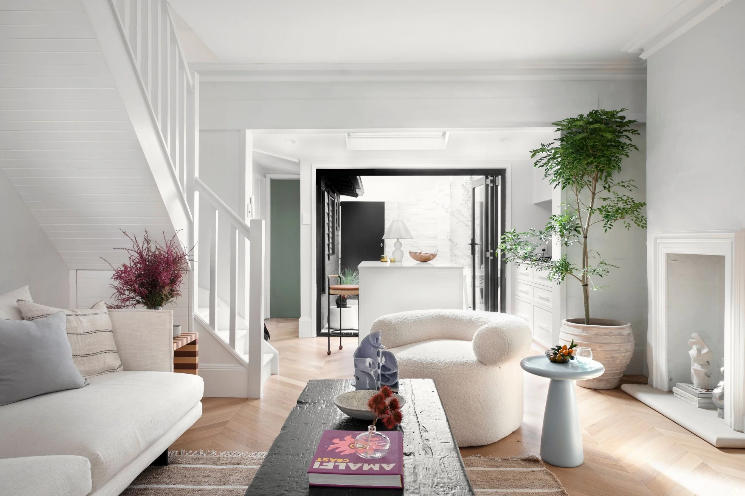 Bright living room with white walls and furniture, including a sofa and a round chair, decorated with plants, pillows, and a black wooden coffee table with a book, colorful desserts, and decorative objects.