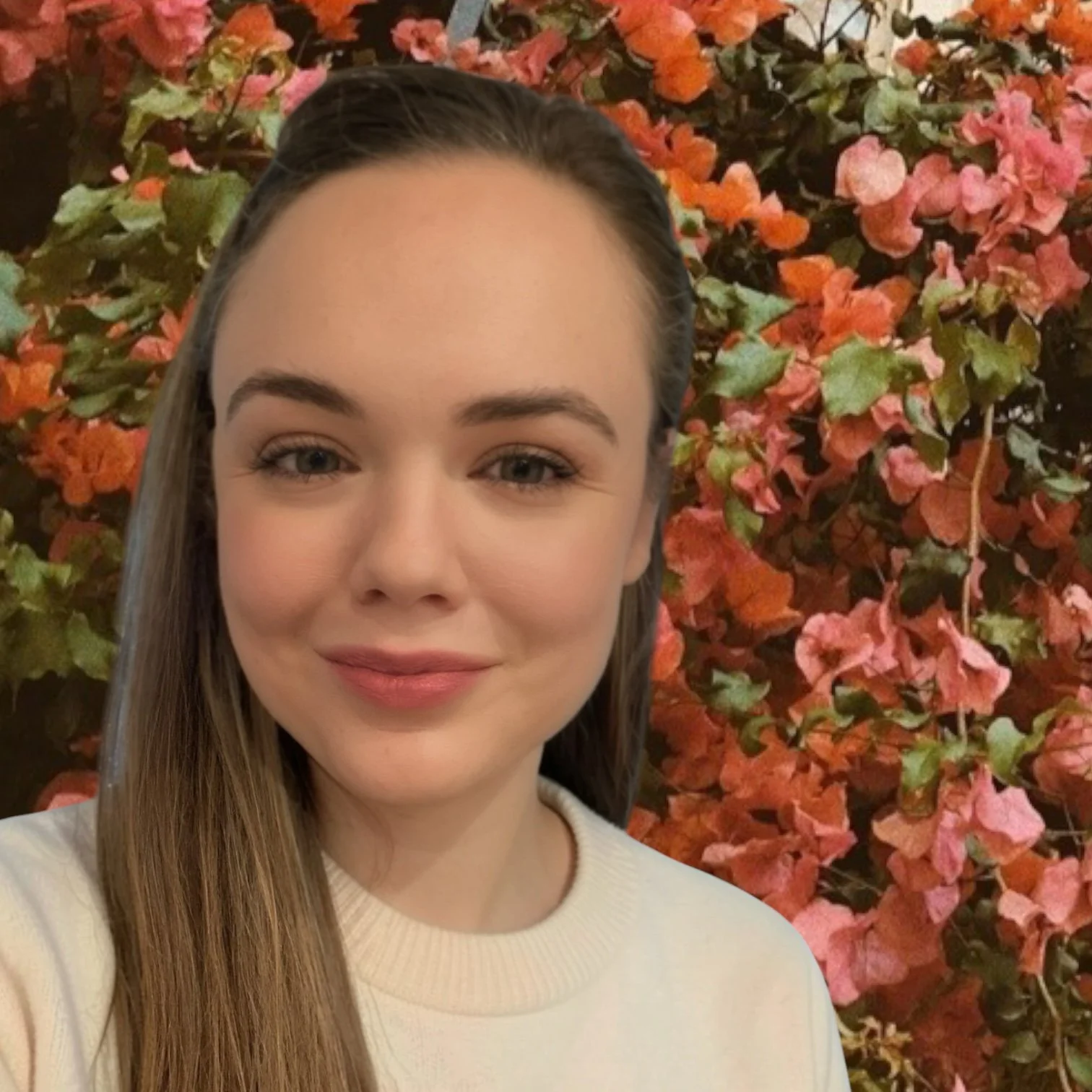 A young woman with straight, long, light brown hair, fair skin, and light makeup, smiling slightly, in front of a bush with pink, orange, and green leaves.