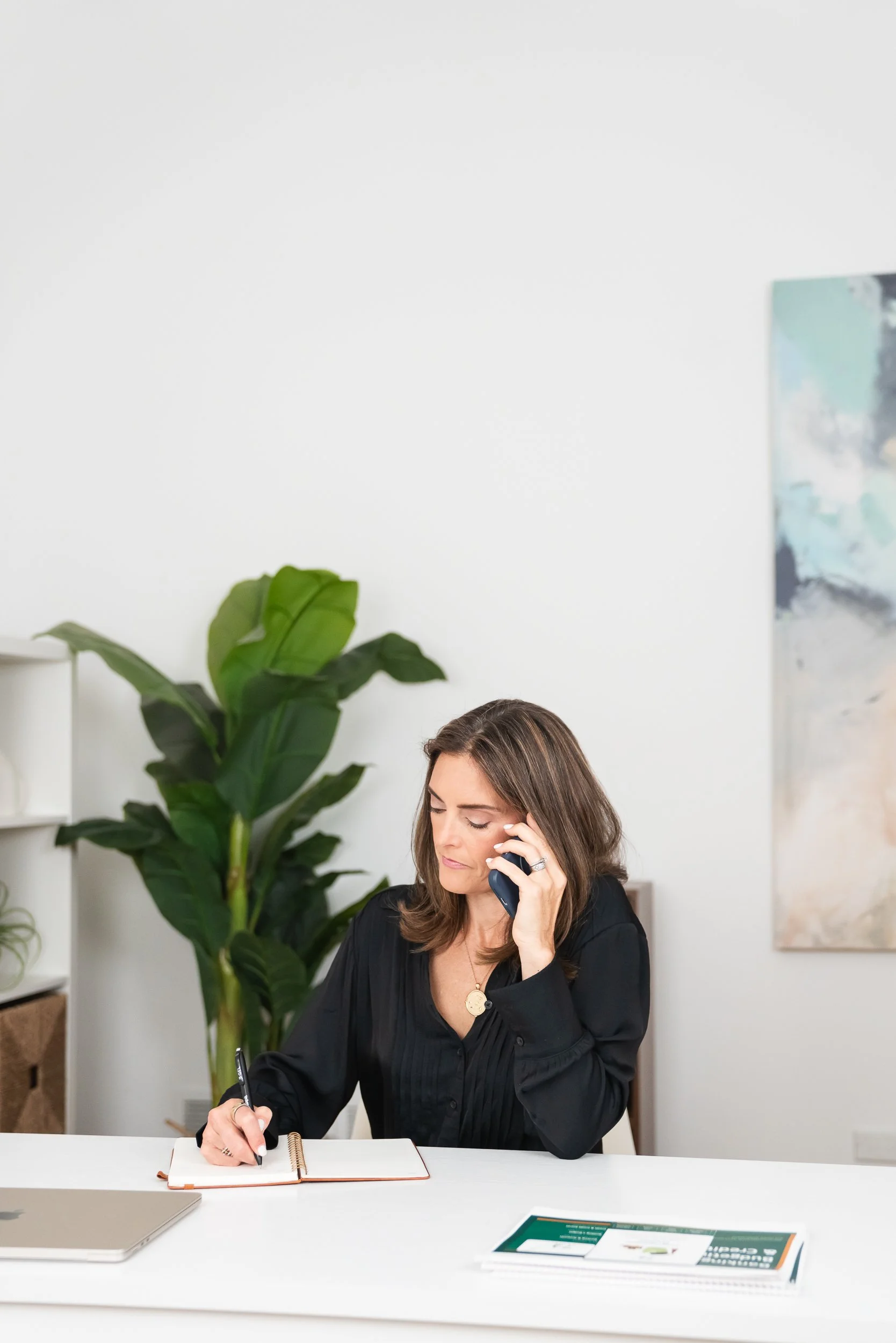 A woman sits at a white desk, talking on the phone, writing in a notebook, with a large green plant behind her and artwork on the wall. There are reading materials and a laptop on the desk.