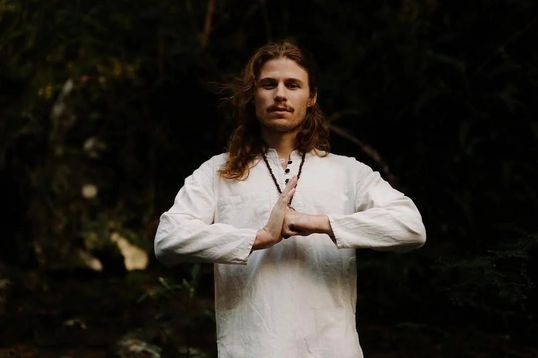 A man with long brown hair and a mustache, wearing a white long-sleeve shirt and a beaded necklace, stands outdoors in a natural setting with dark foliage in the background. He is holding his hands in a prayer position at chest level.