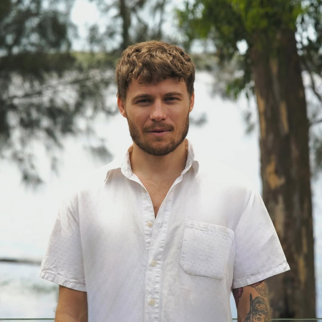 A man with short, curly brown hair and a beard, wearing a white short-sleeve button-up shirt, stands outdoors with trees and a cloudy sky in the background.