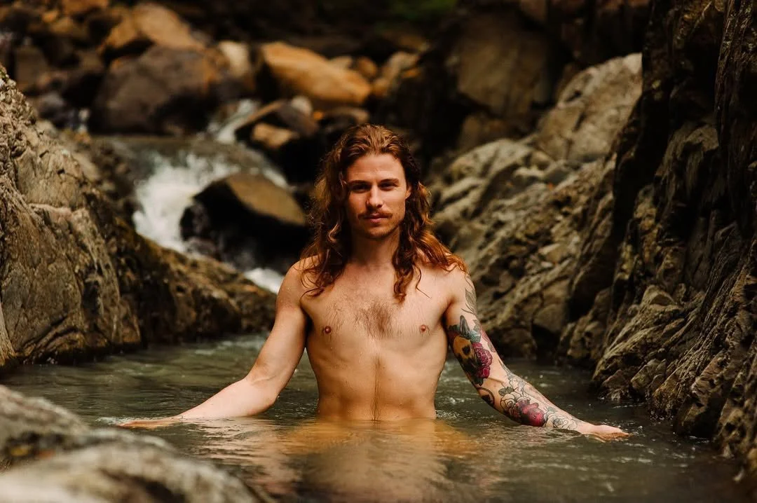 A shirtless man with long hair and tattoos sitting in a rocky creek with water up to his chest.