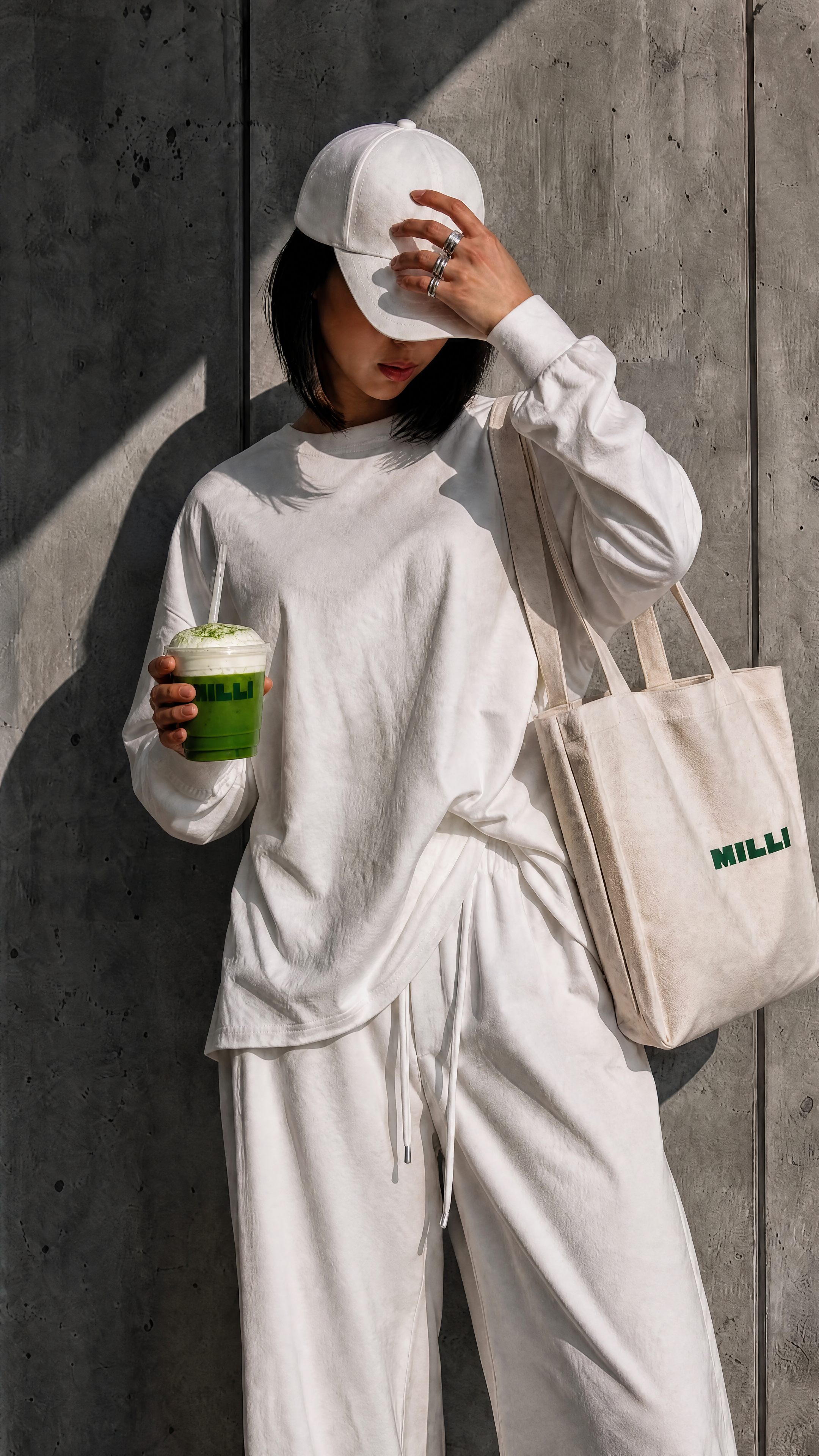 A woman dressed in all white, wearing a white cap and holding a green drink with a straw, standing against a concrete wall. She is carrying a beige tote bag with the word 'MILLI' printed on it and has multiple rings on her fingers.