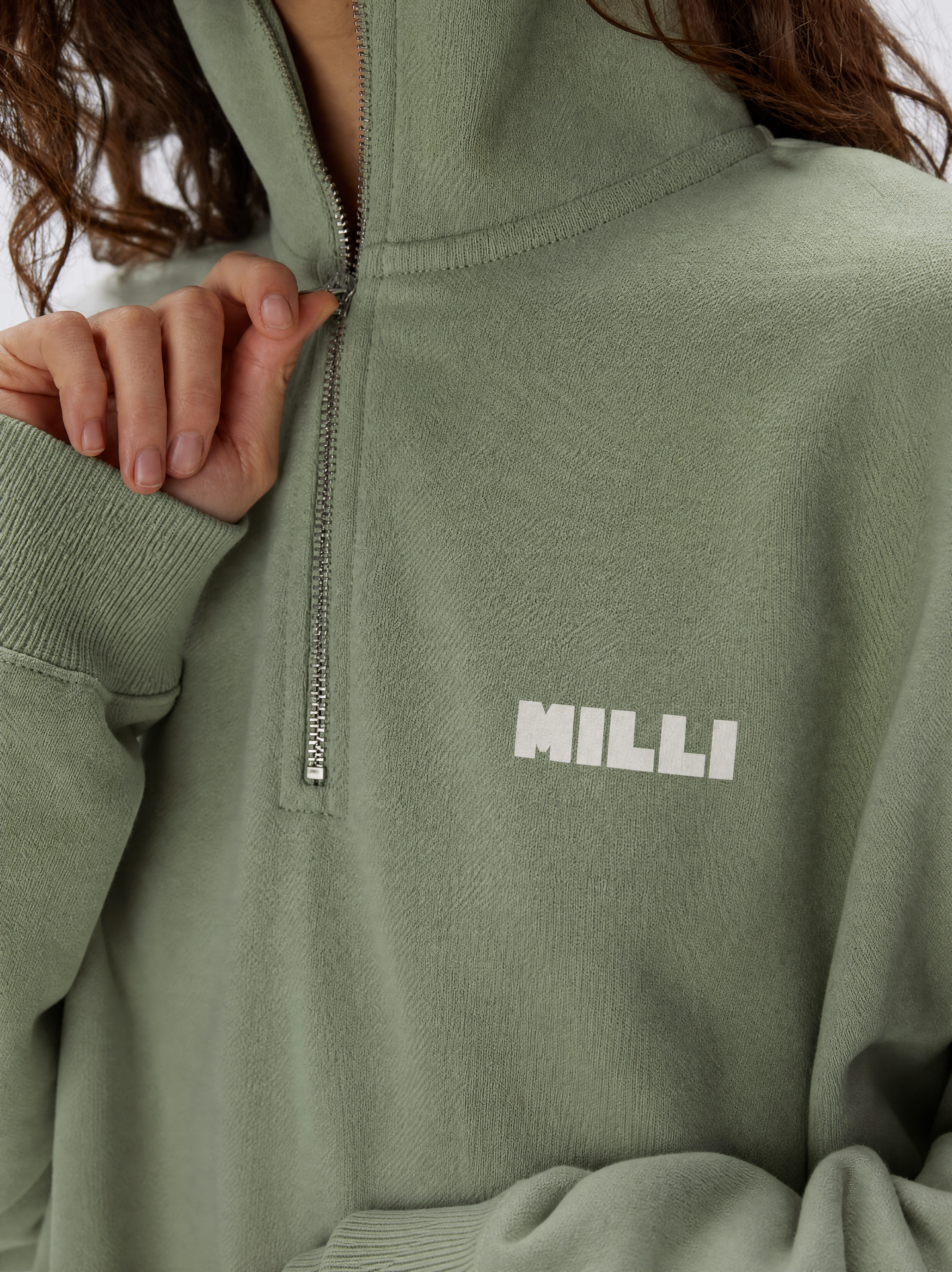A person wearing a green zip-up sweatshirt with the word 'MILLI' printed on it, adjusting the zipper.