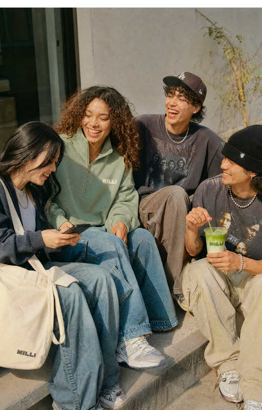 Four young people sitting on steps, smiling and laughing. One woman is looking at her phone, and another person is holding a green drink with a straw. They are dressed casually and appear to be enjoying each other's company.