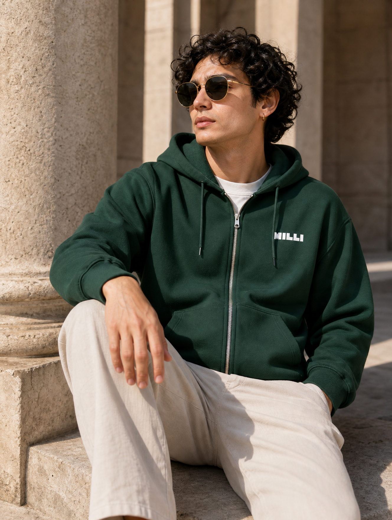 Young man with curly dark hair wearing round sunglasses and earrings, sitting on stone stairs outdoors, dressed in a dark green hoodie and beige pants.