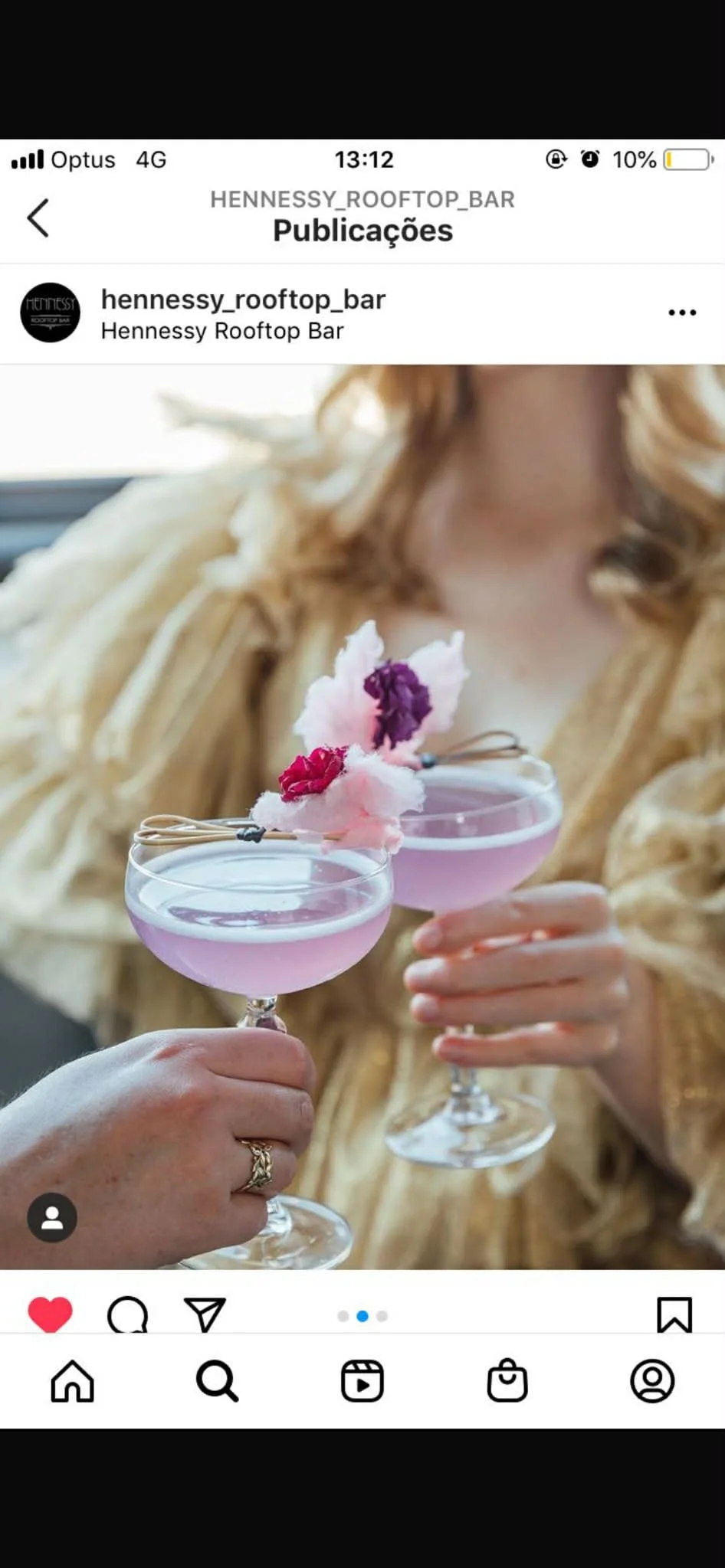 Close-up of two hands holding pink cocktails topped with cotton candy and edible flowers, with a woman in a beige jacket holding the drinks.