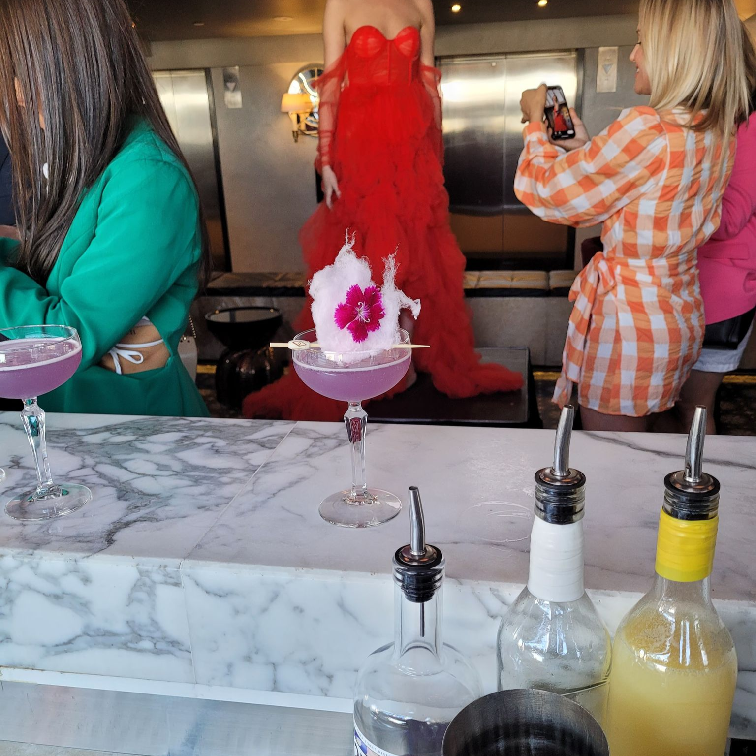A woman in a red dress stands on a stage while several women, some taking photos, observe in a restaurant or bar setting. The foreground shows a marble countertop with three bottles and glasses, and two pink cocktails garnished with a flower and cotton candy.