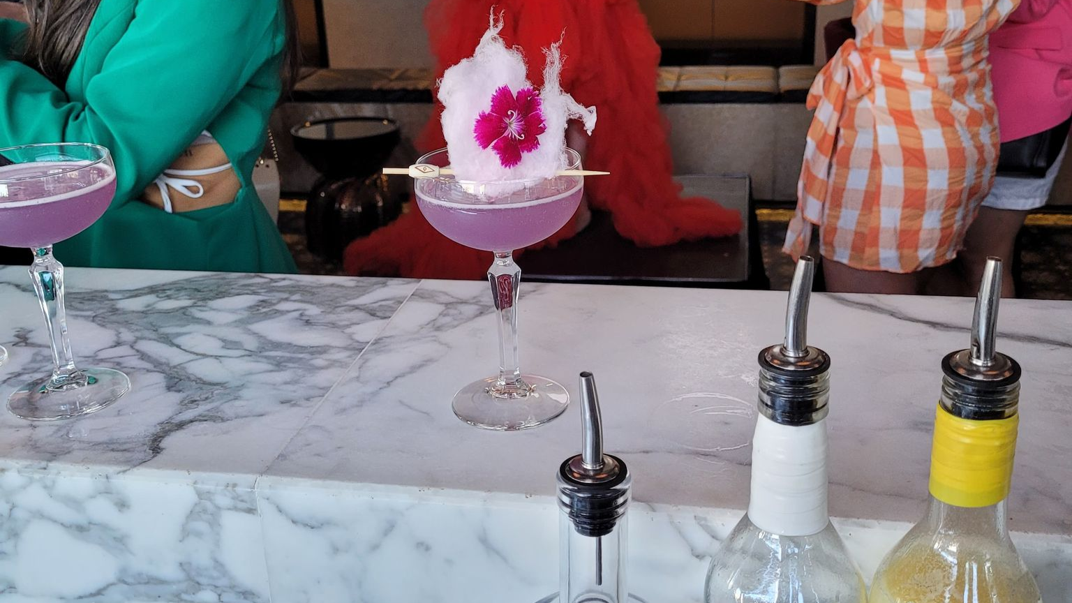 A marble bar counter with three cocktails. The middle one is a pink cocktail garnished with cotton candy and edible flowers. There are condiment bottles with sauce shakers in front of the cocktails. People are standing in the background, including an individual wearing a green jacket and another in orange and white checkered shirt.