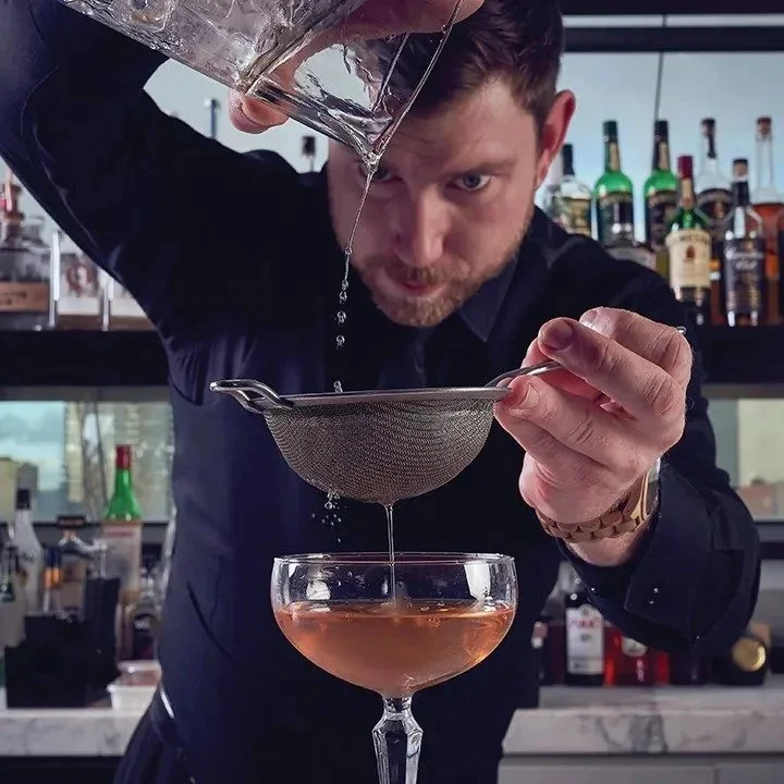 Man pouring a cocktail through a strainer into a glass behind a bar with bottles in the background.