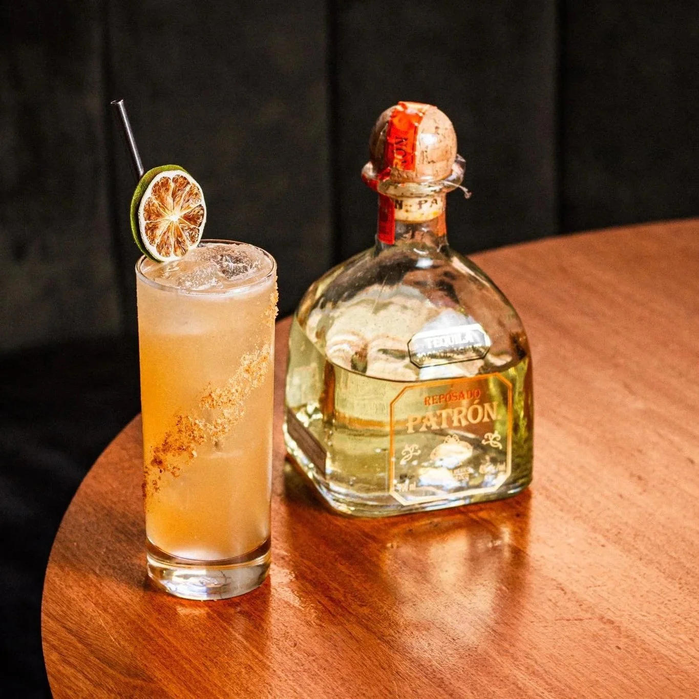 A glass of cocktail with a dried lime slice garnish and a straw, sitting on a wooden table next to a bottle of Patrón tequila.