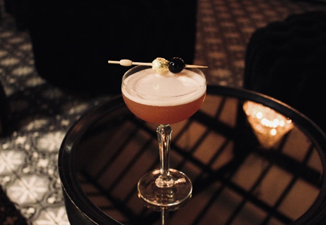 A pink cocktail in a martini glass garnished with a skewer of blackberries and gold dust, on a black round table with a lattice pattern.