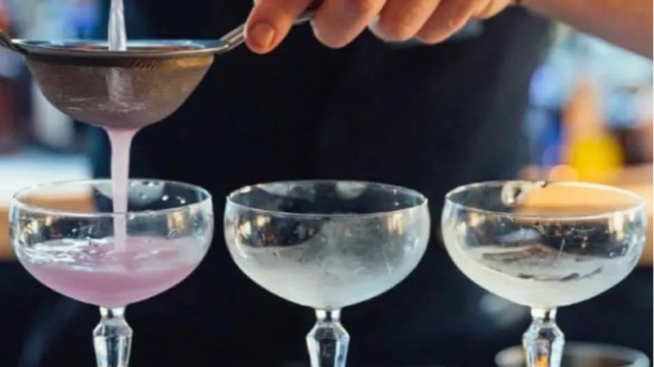 A bartender pours a purple cocktail into a coupe glass while three other coupe glasses are on the bar counter.