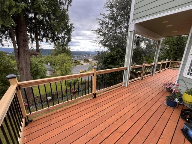 Wooden deck with railing overlooking a suburban neighborhood with trees and houses, cloudy sky in the background, part of a house with siding visible on the right.