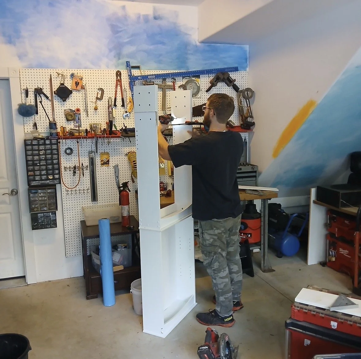 Man working with tools in a garage workshop with pegboard wall holding various tools and equipment.
