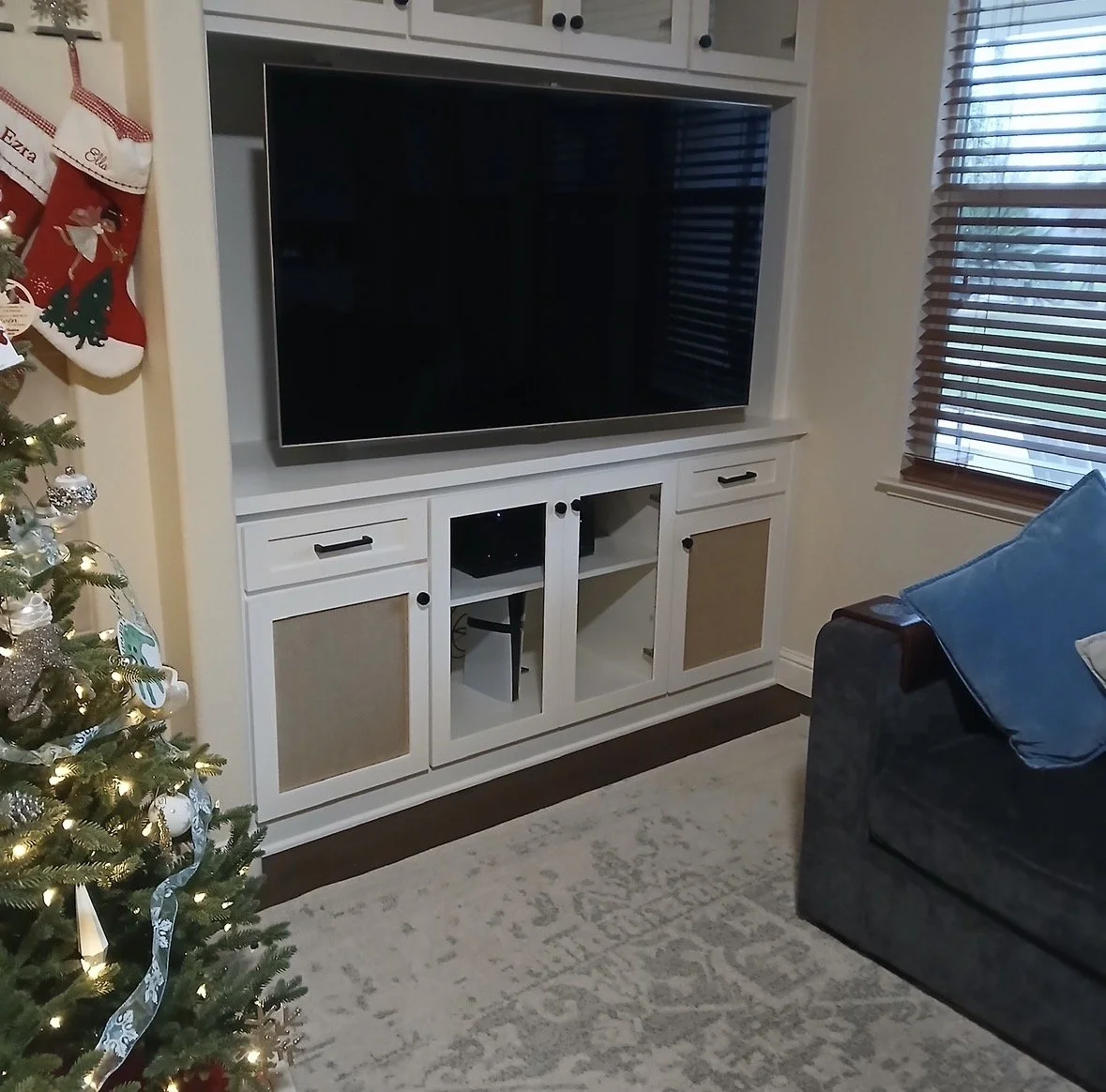 Living room with a TV on a white entertainment center, decorated Christmas tree with lights and ornaments, and a window with blinds.