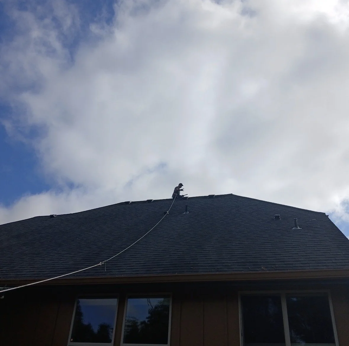 A person sitting on the roof of a house, cleaning or inspecting, with a safety harness attached, under a cloudy sky.