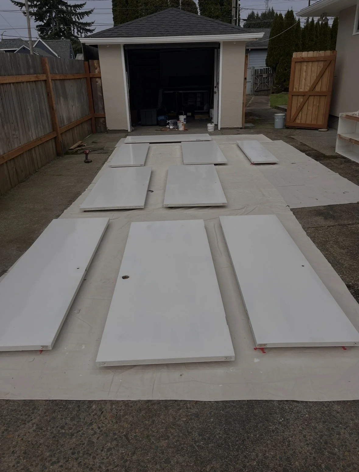 Outdoor backyard scene with white cabinet doors laid out on protective covering, in front of a garage with open door, surrounded by wooden fencing and trees.
