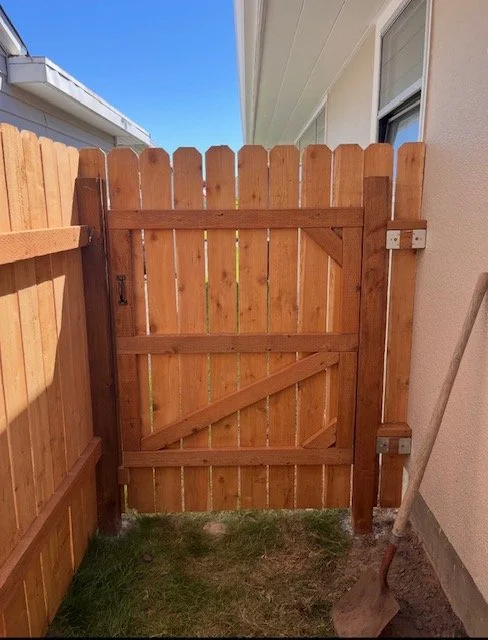 A wooden gate with a latch, part of a fence, next to a house with siding, and a shovel resting nearby on the ground.