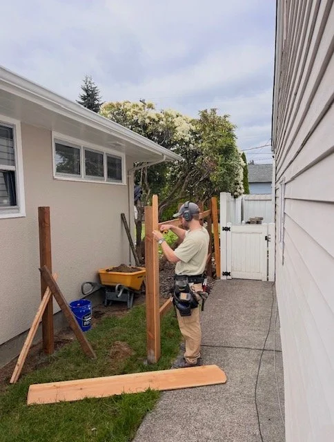 A man wearing a baseball cap, sunglasses, and work gloves building a wooden fence between a house and a shed in a backyard. He is using a power drill on a vertical fence post. There are tools and materials around him, including a yellow wheelbarrow, a blue bucket, and a long piece of wood on the ground. A white gate and trees are visible in the background.
