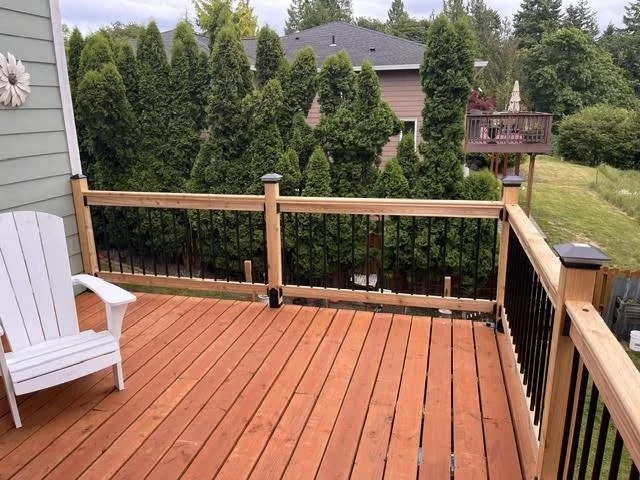 View of a backyard deck with wooden flooring and black and wooden railing, neighboring houses, and green trees.