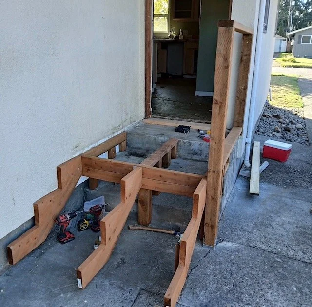Wooden framing for a porch or step being built outside a house, with tools and construction materials around.