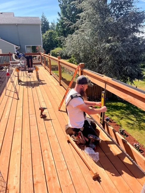 A man working on building or repairing a wooden deck outdoors, kneeling with a level tool, surrounded by construction tools and materials, with trees and a house in the background.