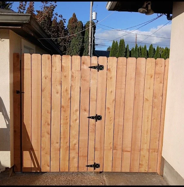 A newly installed wooden gate with black hinges in a backyard, with a clear blue sky and some trees in the background.