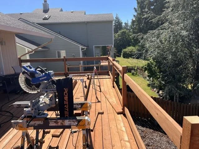 Construction of a wooden deck on the back of a house, with power tools and lumber, in a suburban neighborhood with trees and neighboring houses.