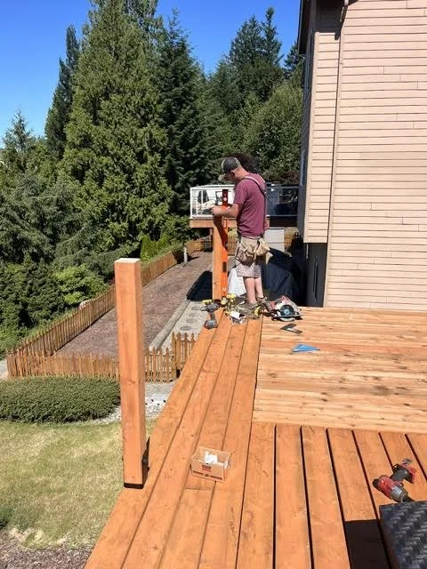 A man working on a house deck, with tools and wood planks, overlooking a backyard with trees and a garden.