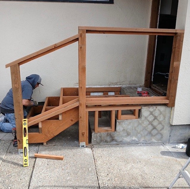 Person installing a wooden staircase or ramp outside a building, with various tools visible on the ground.