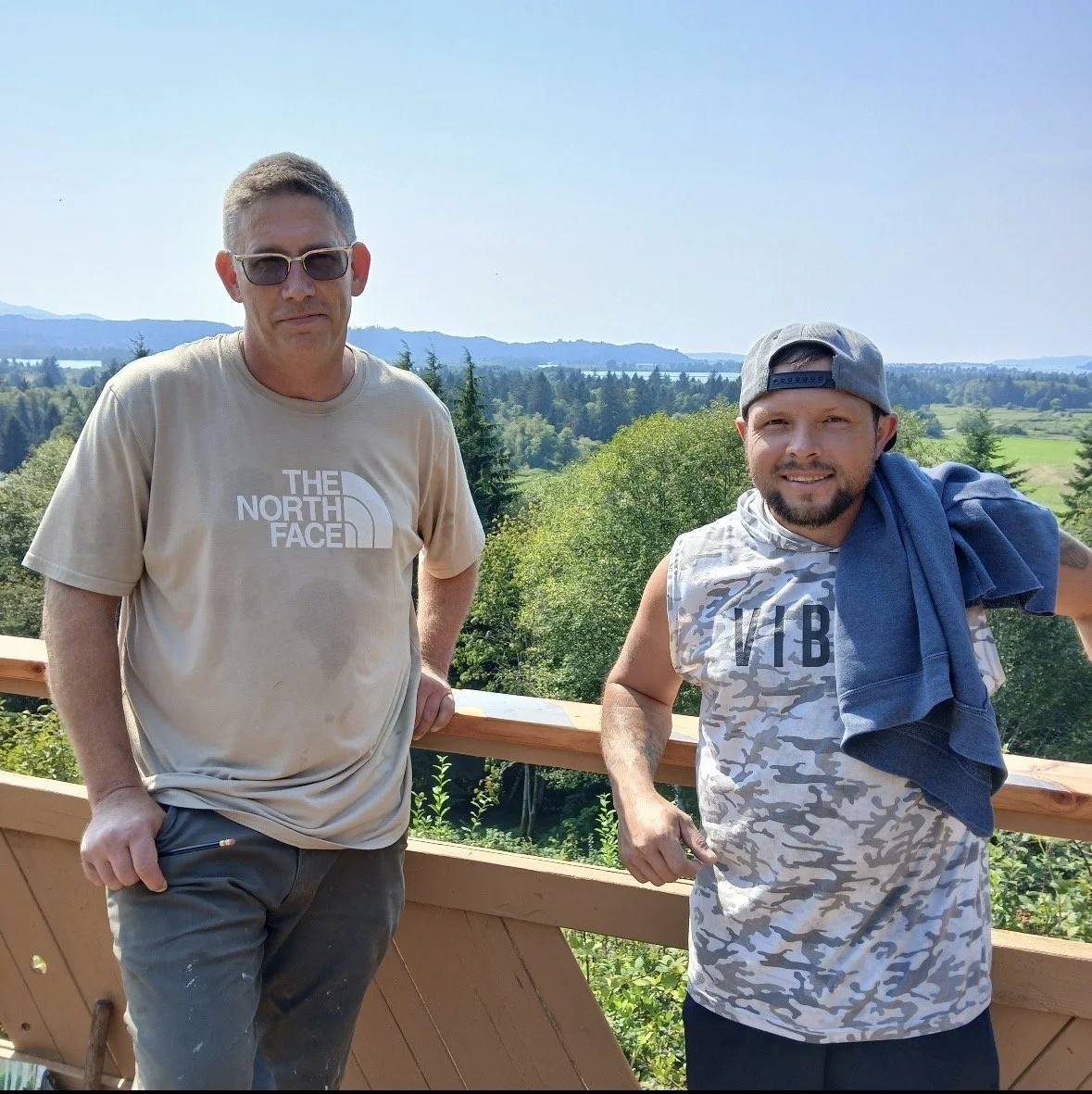 Two men standing outdoors on a wooden balcony with a scenic view of trees, hills, and water in the background on a sunny day, one wearing sunglasses and a beige The North Face t-shirt, the other wearing a gray cap, sleeveless shirt with VIB printed o