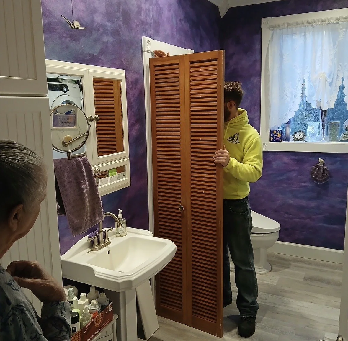 A man in a yellow hoodie stands behind a wooden louvered closet door in a purple bathroom, hiding his face. An elderly woman, partially visible, watches him. The bathroom has a white sink, purple walls, a window with lace curtains, and various toilet