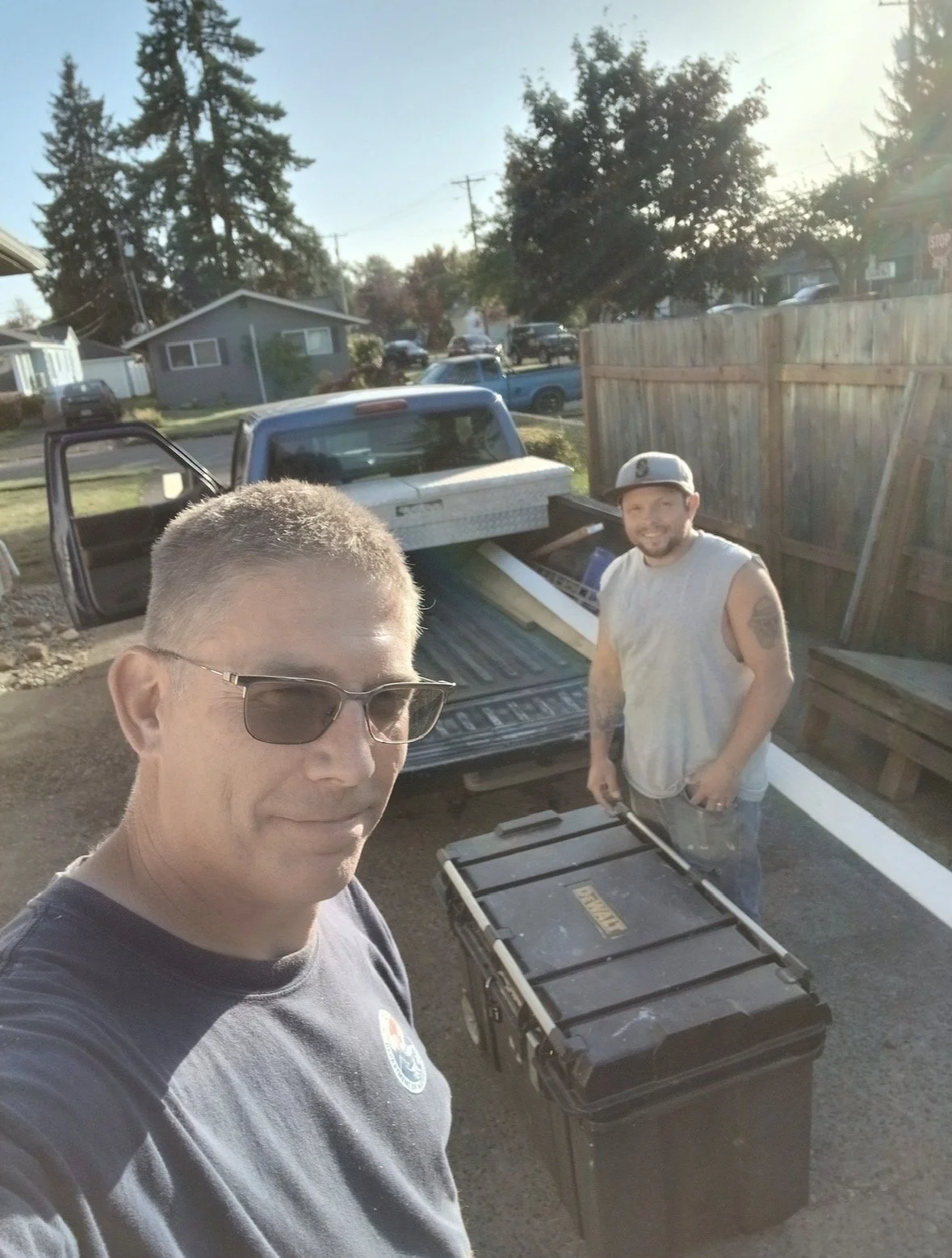Two men taking a selfie outdoors near a pickup truck with an open bed. One man is wearing sunglasses and a t-shirt, the other is wearing a sleeveless shirt and a cap, holding a tool. There is a large plastic storage container near them. The scene is 
