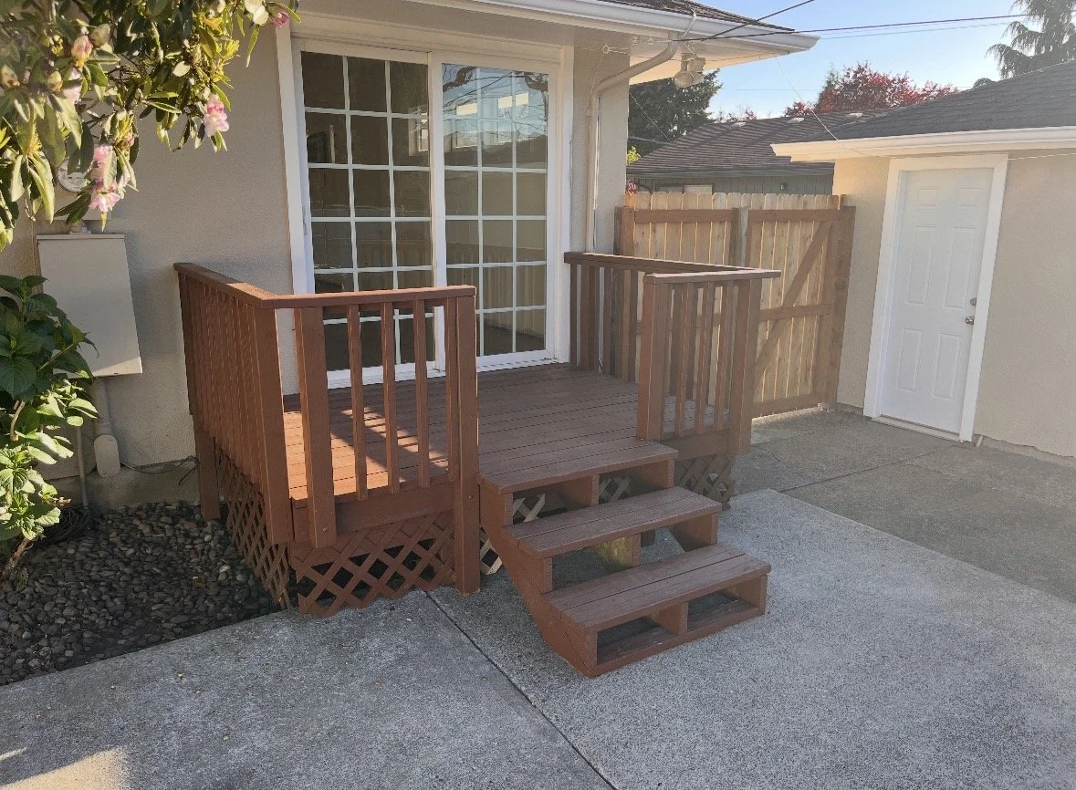 A small wooden deck with stairs and railings outside a house, attached to a sliding glass door. There is a white door on the right and some trees and fences in the background.