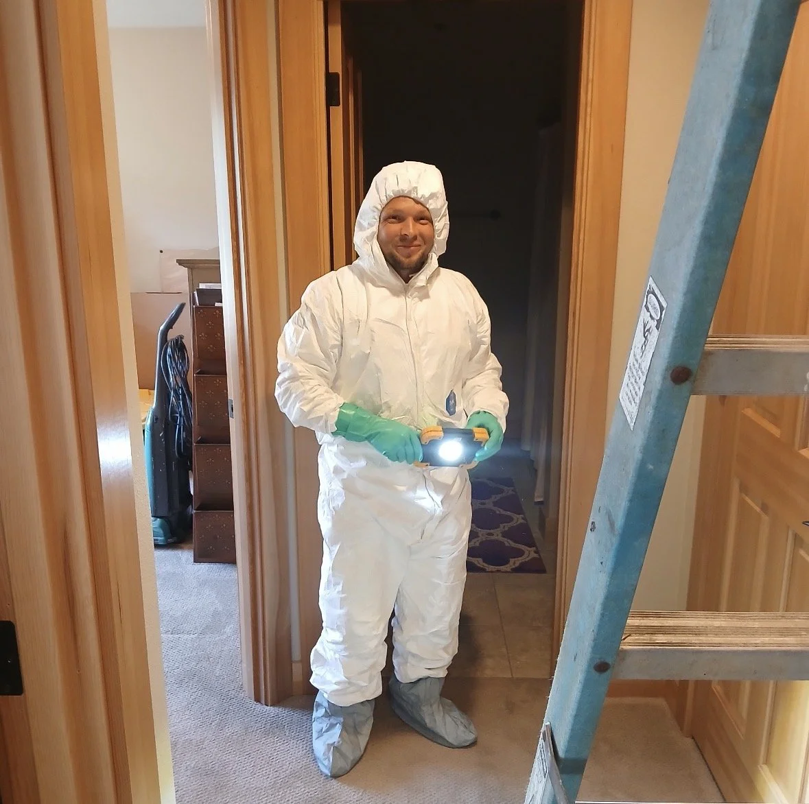 A man in protective white coveralls, green gloves, and overshoes holding a flashlight with a bright light, standing in a doorway inside a house.