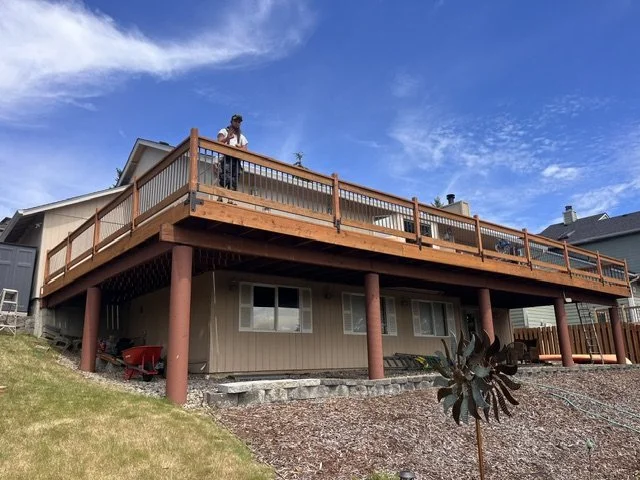 A person stands on a large wooden deck attached to a house, with a clear blue sky in the background.