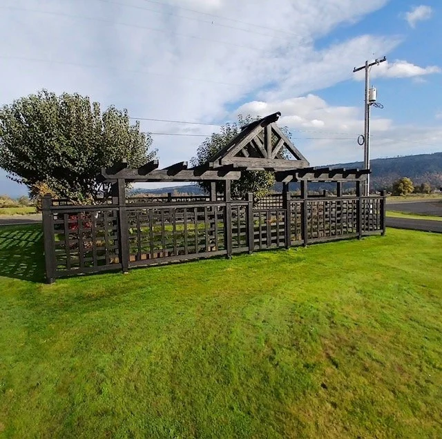 A wooden pergola with a trellis fence surrounding it, situated on a well-maintained grassy lawn, with trees and a utility pole in the background under a partly cloudy sky.