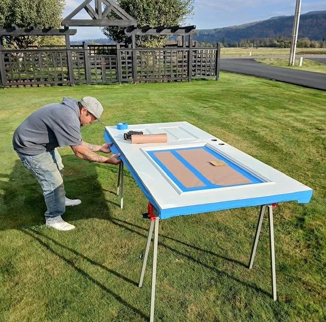 A man working on a door in an outdoor setting, with a table covered in brown paper and tape, likely preparing the door for painting or sealing.
