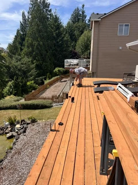 Person installing or working on a wooden deck outside a house on a sunny day with greenery and trees in the background.