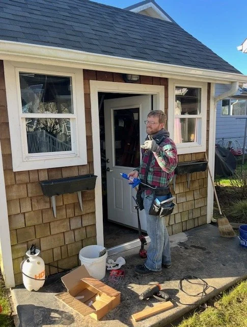 A man with glasses, wearing a plaid shirt and jeans, is standing outside a small house with brown shingles and white trim. He is holding a blue item and a tool, with a tool belt around his waist. There are gardening supplies, a broom, and a cardboard